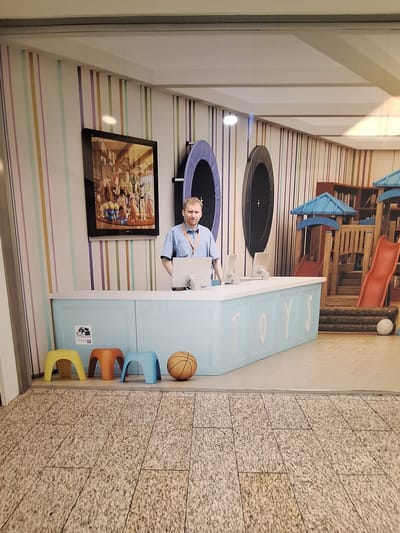 The image depicts an interior scene featuring a man standing behind a light blue counter with "TOYS" written in white on its facade. The man, wearing a light blue shirt and an orange lanyard, is looking towards the camera with a slight smile. On the counter are two computer monitors. To the left of the counter, there are three small, colorful stools in yellow, orange, and blue, and a basketball. Behind the counter, a large poster featuring characters from the movie Toy Story is displayed on a wall with vertical pastel-colored stripes. Further to the right, a wooden playground structure with blue roofs and red slides is visible, along with two large, dark circular objects that appear to be trampolines. The flooring in the foreground is tiled with a speckled gray pattern. There is no indication or discernible information within the image that places the scene in Bochum, Germany.