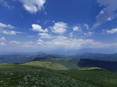 A panoramic, high-angle view captures a verdant, rolling mountain landscape under a bright blue sky dotted with white clouds. In the foreground, a grassy slope is covered with scattered green vegetation. The mid-ground reveals more undulating green hills, some with scattered trees and a patch of bright green grass catching the sunlight. Shadows cast by unseen clouds create areas of darker green and deep shadow on the slopes. The background consists of layered mountain ranges receding into the distance, their muted blue tones fading into a hazy atmosphere. A faint suggestion of a road or track is visible on one of the distant hillsides. The scene is devoid of any people or man-made structures, presenting a natural, expansive vista. This vista is identified as being in the vicinity of Brestovo, Bulgaria.
