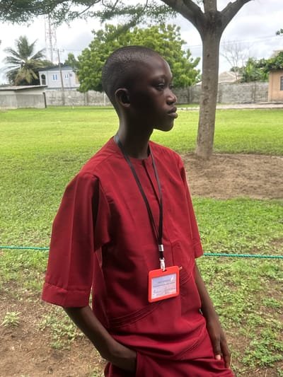 A young Black individual, appearing to be male, stands outdoors wearing a dark red, short-sleeved tunic with a high neckline and subtle decorative stitching on the chest. A black lanyard with a white clip is around his neck, holding a bright orange identification card with text and handwriting. His left hand is tucked into the pocket of his tunic. He has a short buzz cut and is looking to his right with a neutral expression. The background consists of a lush green lawn with scattered patches of dry earth and a thin green rope fence running horizontally across the lower portion of the frame. A large tree trunk is visible to the right of the individual. In the distance, beyond the lawn, there are buildings, including one with white trim and blue windows, and a tall radio tower. The sky is overcast. The overall setting appears to be a school or community grounds in Itoga, Nigeria.