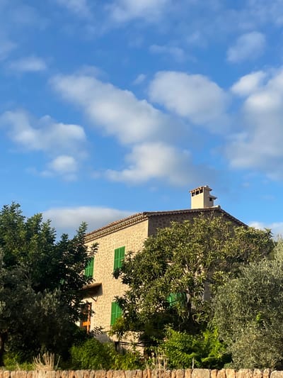 A traditional stone house is depicted from a low angle under a blue sky with scattered white clouds. The structure, located in Deià, Spain, features a light brown, rough-hewn stone facade. Multiple rectangular windows, each fitted with closed green louvered shutters, are visible on at least two stories. A terracotta tiled roof caps the building, from which a distinctive, multi-tiered, light-colored chimney rises on the right side. Dense green foliage, including trees with varying leaf densities and what appears to be an olive tree on the right, partially obscures the house. A reddish-brown door is partially visible on the left side of the house. In the immediate foreground, a dry-stone wall constructed of light-colored, irregular stones spans the width of the frame. No individuals, animals, or active interactions are present in the scene. The environment is well-lit by natural sunlight.