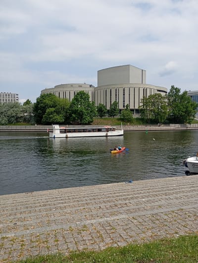 The image captures a waterfront scene in Bydgoszcz, Poland, likely on the Brda River. In the background is the Opera Nova building, a modern architectural structure with a distinct cylindrical and cuboid design. A white tour boat is moving along the river, its white facade contrasting with the dark water. Closer to the foreground is a brightly colored kayak with two occupants. Further on, a white sailboat is moored. The riverbank is composed of stone steps leading down to the water's edge, with grass and vegetation visible at the bottom. Pedestrians are strolling along the river embankment. The sky is overcast, but the scene overall appears to be taken on a bright, summer day.