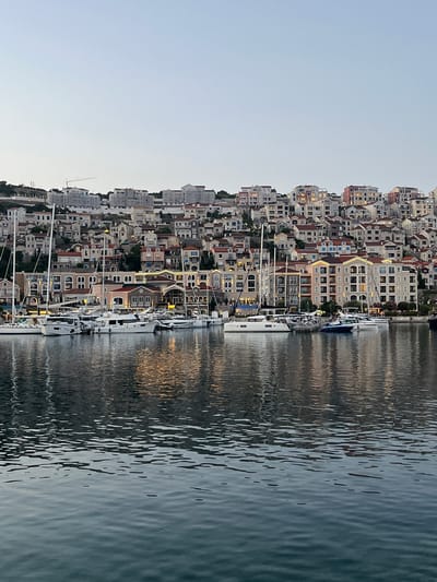 The image depicts a waterfront scene in Gošići, Montenegro, featuring a marina filled with various yachts and sailboats. In the background, a hillside is densely populated with multi-story buildings, many of which are illuminated, suggesting it is dusk or early evening. The architecture of the buildings appears to be Mediterranean-inspired, with tiled roofs and light-colored facades. The water in the foreground is calm, reflecting the lights of the buildings and the boats. The sky is clear and pale, indicating the transition from day to night. There are no people visible in the image.
