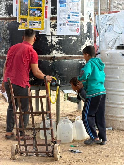 An outdoor scene in a-Zawayda, Palestinian Territory, features three individuals collecting water from a large, dark-colored plastic water tank. The tank, heavily weathered with peeling material, is mounted on a metal stand and has multiple faucets. Affixed to its surface are a yellow "Save the Children" poster depicting safety instructions and a white informational poster bearing the "UNICEF" logo, with visible Arabic text.

An adult male, viewed from behind, wears a red short-sleeved shirt and dark trousers. He manipulates a yellow hose connected to one of the tank's faucets, appearing to control the water flow. Adjacent to him, a younger child, dressed in a grey long-sleeved shirt, crouches and holds a small, patterned plastic cup to collect water actively flowing from the hose. Standing nearby is an older child, wearing a teal hooded sweatshirt with grey elbow patches and dark blue track pants with light blue stripes.

In the foreground, a rusty, two-wheeled metal hand truck is visible, carrying one clear plastic jerrycan filled with water. On the sandy ground near the children, three additional clear plastic jerrycans are present, one of which has Arabic writing ("أبو أحمد") inscribed on it. Small pieces of debris are scattered on the sandy terrain. To the right, a white plastic water tank and a large white tarp structure are partially visible in the background.