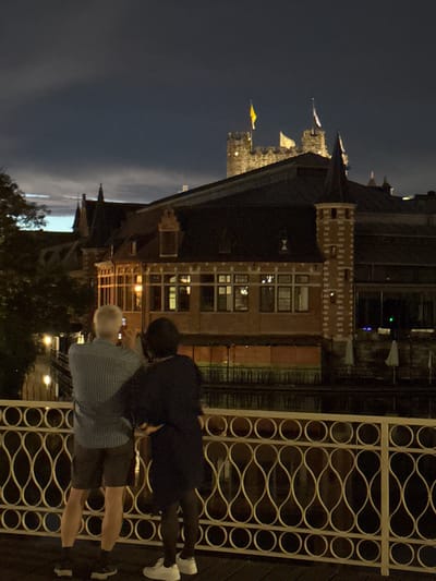Two individuals, a man and a woman, stand on a bridge at night, facing a canal and buildings. The man, in a striped shirt and shorts, holds a phone, appearing to take a photograph. The woman, in a dark dress and tights, stands beside him, her arm around his shoulder. Behind them, illuminated by internal lights, is a building with large windows and a prominent turret. In the distance, a castle-like structure with flags is visible against a dark sky. The scene is located in Ghent, Belgium, likely near the Gravensteen castle and the canals of the city.