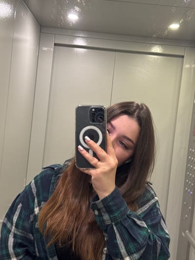A young woman with long, brown hair takes a mirror selfie inside an elevator. She is wearing a dark green and blue plaid shirt over a black top. Her left hand, holding her phone, is visible, along with her right hand, which is partially obscuring her face. The elevator walls appear to be metallic, and a panel with buttons is visible to the right. The lighting is bright and seems to be coming from overhead. The location is identified as the city of Сокол, Russia.