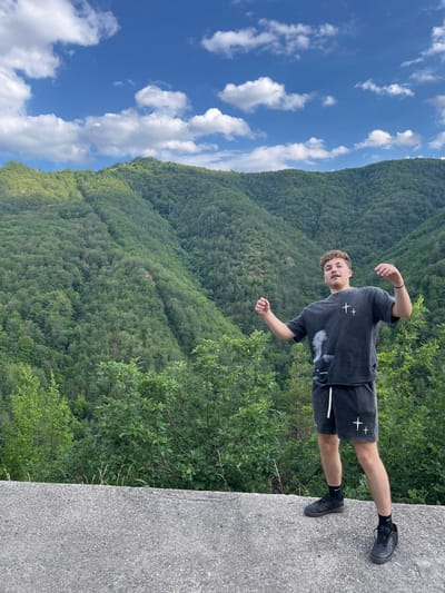 A young man is standing on a concrete ledge outdoors. He is wearing a dark grey t-shirt with white crosses and a graphic print, and matching dark grey shorts with white crosses. He has black socks and black sneakers. The man is looking towards the camera with his arms slightly raised and hands open. Behind him, a lush green, heavily forested mountain range stretches into the distance under a bright blue sky with scattered white clouds. The setting appears to be a natural, mountainous landscape. The location is not specified as being in the city of Pechinska, Bulgaria.