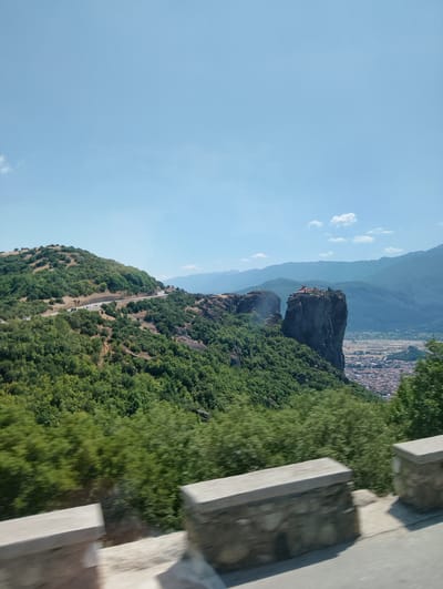 This is a landscape photograph taken from a moving vehicle, capturing a scenic view of the Meteora rock formations and the town of Kalambaka, Greece. The foreground features a blurred roadside barrier constructed from stone and concrete, indicating the vantage point of the photographer. Beyond the barrier, a winding road is visible, with several cars driving on it, suggesting it's a public route. The road cuts through a lush green hillside covered in dense vegetation and trees. Dominating the midground are the iconic sandstone pillars of Meteora, one of which supports a small monastery complex with red-tiled roofs. In the background, the town of Kalambaka is spread out in the valley, with numerous red-tiled roofs indicative of residential buildings. Further in the distance, a range of hazy blue mountains forms the horizon under a clear, pale blue sky with scattered white clouds. The overall impression is one of a bright, sunny day offering a panoramic view of the historical and natural landscape.