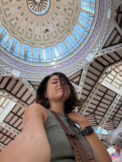 This is a selfie taken from a low angle, looking up at the dome ceiling of the Central Market of Valencia, Spain, a historic building located in the city of Alboraia/Alboraya. The woman in the foreground is looking towards the camera with a slight smile. Her dark hair is slightly windswept across her face. She is wearing a green ribbed tank top and has a brown woven bag slung over her shoulder. On her left wrist, she wears a smartwatch displaying the time as 10:44 AM and the date as 17 H E P B. The background features the intricate architecture of the market's dome, with its stained-glass windows allowing natural light to filter through, illuminating the decorative ceiling with floral motifs and geometric patterns. Structural beams and supports of the market are also visible. To the right of the woman, a portion of an elderly woman with glasses is visible, appearing to look at something out of frame.
