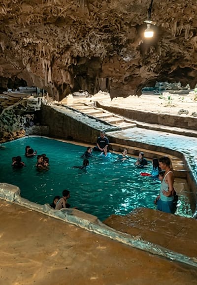 A group of people are bathing in a turquoise swimming pool located inside a cave. The cave ceiling is adorned with stalactites, and a light fixture hangs from above. Stone steps lead down to the pool on the right side of the image, and there appears to be another set of steps on the left. Some of the people are in the water, while others are sitting on the edges or stairs. The pool area is surrounded by rocky formations and sand. The image shows a recreational scene within a natural cave environment. The specific location in Penungkiren, Indonesia is not visually discernible from the image itself, but the scene depicts an interior cave pool.