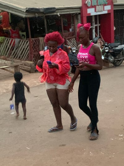 The image depicts an outdoor street scene in Otuocha, Nigeria, featuring three individuals: two adult women and a child. The lighting suggests it is daytime, possibly overcast.
In the foreground, a young child dressed in a dark outfit stands with their back to the camera. Behind them, a woman is seen wearing a bright red, flowing blouse over light-colored striped shorts, with light blue flip-flops on her feet. She holds a smartphone with white wired earbuds, seemingly engaged with the device. Her hair is styled in a distinctive red, curly or braided fashion.
Adjacent to her, another woman stands wearing a hot pink tank top, black leggings, and dark sandals. The pink top has white text printed on it, which clearly reads "DRINK LOVE RELAX". Her hair also appears to be styled or wrapped in pink. Her left hand is positioned near the arm of the woman in red.
In the background, a red building with a partial white sign (text illegible) is visible, along with what appears to be a parked motorbike or scooter. Other building structures with metal gates or grates are also present, indicating a commercial or residential area. The ground beneath the individuals is a light, dusty surface, likely dirt or concrete. The overall atmosphere is that of a casual, everyday moment in a local setting.