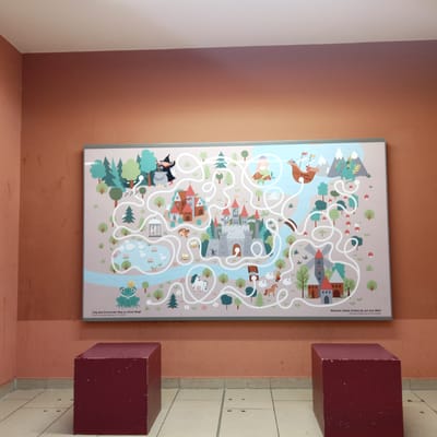 This image shows a maze poster on an orange wall in a public indoor space. The poster depicts a fantasy scene with castles, a witch, a dragon, a mermaid, a ship, a windmill, a frog with a crown, a unicorn, swans, sheep, horses, and carts. The maze is depicted as a winding white path through the scene. Text in German and English is present at the bottom of the poster. Two maroon cube-shaped benches are positioned in front of the poster, one on the left and one on the right. The floor is tiled. The environment appears to be a waiting area or an educational space. This poster, featuring a maze titled "Show the frog the right way to his castle!", is located in the Wartburg Castle in Eisenach, Germany, likely in an area accessible to visitors.
