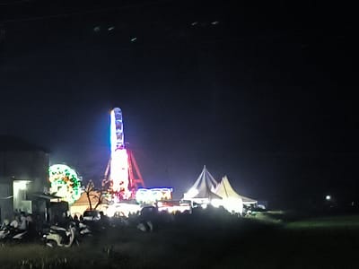 At night in Blangbitra, Indonesia, a fair is illuminated with various rides and tents. A Ferris wheel and a tower ride are prominent features. People are gathered near the rides, and several motorbikes are parked nearby. A building is visible on the left side of the image. The environment consists of dark sky, grass, and open space around the fair.