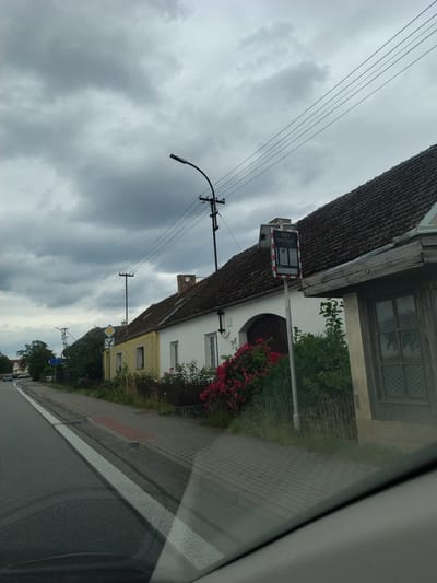 This image captures a street scene in Svatonice, Czechia, under an overcast sky. The photograph is taken from a vehicle, as indicated by the reflection on the right side of the frame.
The primary subjects are buildings and infrastructure. On the left, a traditional building with a dark, textured roof dominates the foreground. A street sign with text and a digital display is mounted on a pole near the building. The sign appears to be a speed display, with the text "Vaše rychlost:" (Your speed:) and a digital readout. Adjacent to this building, another structure with yellow and white facades is visible, with red flowers blooming in front of it. A sidewalk made of brick pavers runs alongside the buildings.
The scene is set on a road, with a grey asphalt surface and white lane markings visible. Several utility poles and power lines stretch across the frame, suggesting an inhabited area. Trees are also present, contributing to the somewhat rural or suburban feel of the location.
The weather appears to be cloudy and possibly damp, given the muted light and the appearance of the sky. The time of day is not clearly discernible, but the lighting suggests it is daytime. No people are visible in the frame, and the overall mood is neutral, focusing on the environment.
The visible text on the sign is "Vaše rychlost:".