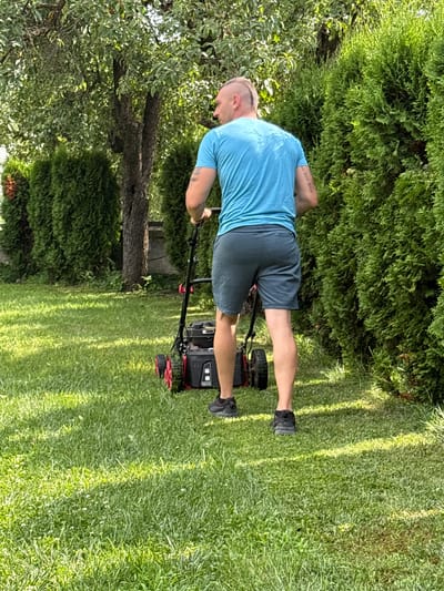 A man with a mohawk haircut is mowing a lawn with a red and black lawnmower in a residential backyard. He is wearing a blue t-shirt and gray shorts, and dark sneakers. The lawn is green and well-maintained, with patches of clover visible. Tall evergreen hedges line the right side of the yard, and a large tree with green leaves is in the background. The scene appears to be in Oreshak, Bulgaria, given the context provided.
