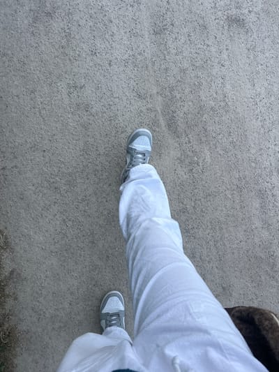 A downward-angled shot depicts a person's lower body, specifically the legs and feet, on a gray gravel surface. The individual is wearing light gray sneakers and loose-fitting white pants. The setting appears to be an outdoor area with a gravel ground. The environment suggests an urban setting, such as a park path or sidewalk. There is no information present within the image to suggest that the location is specifically in Marquette-lez-Lille, France.
