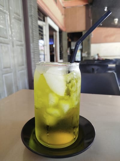 A tall glass of iced green tea with a black straw sits on a black saucer on a beige table. The glass is sweating, indicating the drink is cold. Ice cubes are visible within the green liquid. In the background, the blurry facade of a building is visible with windows and what appears to be a covered walkway. The scene suggests an outdoor seating area of a cafe or restaurant in Blangmatangkuli, Indonesia, given the architectural style. No people are clearly visible in the frame.