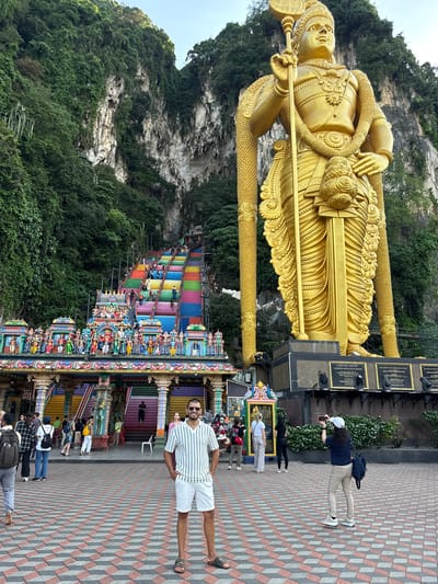 The vibrant image depicts a lively scene at the iconic Batu Caves, situated in the Selayang Municipal Council, Malaysia. Dominating the left side is the colossal golden statue of Lord Murugan, intricately detailed and holding a sacred spear (Vel). The statue stands proudly against a backdrop of towering, verdant limestone karsts.

To the right of the statue, a long and steep flight of stairs, vividly painted in rainbow colors, ascends towards the cave entrance. Many visitors are seen climbing or descending these steps, contributing to the bustling activity. At the foot of the stairs, several ornate temple structures and small vendor stalls are visible, adding to the cultural ambiance.

In the immediate right foreground, a man wearing a striped white and green shirt, white shorts, and sunglasses stands facing the camera with a smile, posing on a distinctive red, white, and grey patterned paved ground. Other people are scattered throughout the scene; some are observing the golden statue, while others are near the base of the stairs or making their ascent. One individual is captured taking a photo near the statue's base.

The scene is bathed in bright daylight, possibly under an overcast sky, highlighting the rich colors and details. The overall impression is that of a popular tourist and religious site. Dark plaques with gold framing are visible at the base of the golden statue, but any text on them is illegible.