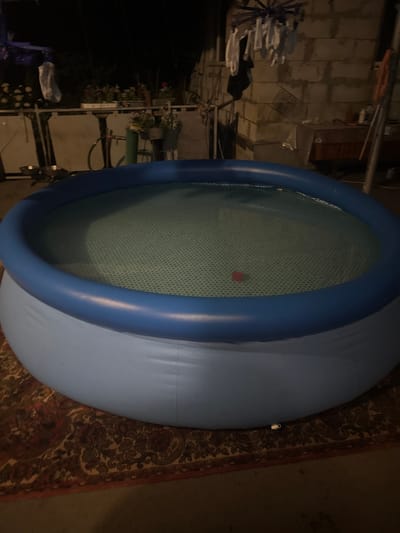 An inflatable swimming pool filled with water sits on a patterned rug in an outdoor area at night. The pool has a blue inflatable ring and a light blue base. The water is clear and has a patterned bottom. A small red object is visible in the water. In the background, there is a concrete wall, a clothesline with laundry, some potted plants, and a table. There are no people visible in the image. The location is Shakhty, Russia.