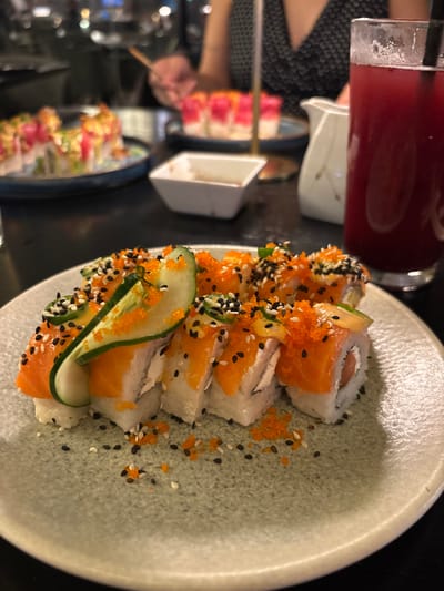 Sushi rolls fill a textured plate. The rolls feature salmon, rice, and what appears to be cream cheese. They are topped with cucumber, sesame seeds, and orange tobiko roe. A red beverage in a glass sits to the upper left. Other dishes are visible on the table in the background, including a small white bowl and another plate of sushi. The lighting in the restaurant is subdued.