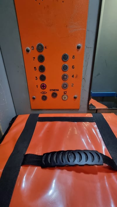 The image shows the control panel of an elevator with numbered buttons from 1 to 9. Button 1 is illuminated in red. Below the numbered buttons are control buttons with symbols for up and down arrows, a cancel button labeled "ОТМЕНА", and a button with a bell symbol. An orange bag with black straps and a black handle is placed in front of the elevator panel. The elevator panel is orange with grey surrounding elements. The background shows a reflection in a metallic surface and part of a blue railing. The image is taken from a slightly low angle, looking up at the control panel.