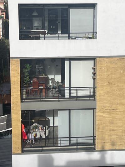 This image depicts a multi-story modern building facade located in the city of Bogota, Capital District, Colombia, viewed from a slightly elevated position. The building features a predominantly white exterior with large, dark-framed windows and balconies on each visible floor. A section of the right side of the building is clad in light yellow brick.

**Visible Floors (from top to bottom):**

1.  **Top Floor:** This level displays an interior space appearing to be a kitchen or dining area. Visible elements include a white pendant lamp, a white cabinet or refrigerator, a dark counter or table, and a framed picture on the rear wall. A small plant is discernible on the exterior balcony ledge.
2.  **Middle Floor:** This floor shows a living or dining area with a large fiddle-leaf fig plant positioned on the left. A wooden dining table with matching chairs is centrally located. To the right, an armchair and a black, rounded object, possibly a pet bed, are visible, with a small light-colored object near the base of the black object, possibly a small animal or decor. The balcony features a black metal railing. On the exterior yellow brick wall to the right of this balcony, three small, rectangular, tiered shelves are affixed.
3.  **Bottom Floor:** This floor also reveals an interior space, likely a living or dining area. A white table with white chairs is present. To the left of the table, a compact drying rack or similar structure holds colorful fabric items, including red, yellow, blue, and green pieces. Small potted plants are arranged on the table. The balcony is equipped with a black metal railing.

No individuals are visible within the building or on the balconies. The overall scene suggests a residential building in an urban environment, with glimpses of other buildings and foliage in the background to the left.