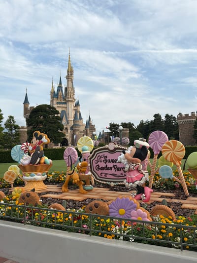 The image depicts a whimsical outdoor scene in Urayasu, Japan, likely at Tokyo Disneyland, characterized by a fantasy-themed display. In the background, a grand castle with multiple spires and ornate architecture dominates the skyline under a partially cloudy sky. In the foreground, a festive arrangement of oversized candy-themed decorations is prominently displayed.

Central to the scene are large, colorful lollipops in various swirling patterns and hues, including pink, purple, orange, and yellow. Accompanying these are character figures: Mickey Mouse is depicted within a giant ice cream sundae-like structure, holding a candy cane and surrounded by cookies and ice cream decorations. Minnie Mouse, dressed in a pink outfit with a matching hat, stands next to a sign that reads "Sweet Garden Party" in elegant script. Pluto is also present, a cheerful figure holding a small basket.

The display is further enhanced by large, stylized cookies, and flower-like decorations in vibrant colors like purple and pink. The entire setup is set against a backdrop of manicured green lawns and bordered by a low grey wall and a metal fence, behind which a multitude of small, colorful flowers, predominantly yellow and white, bloom in clusters. Trees form a dense line behind the display, and additional castle-like structures are visible further in the distance, adding to the park's fairytale atmosphere. The overall impression is one of joyful celebration and enchantment.