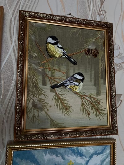 The image shows two framed embroidered pictures of birds hanging on a textured wallpaper. The top picture, which is the primary subject, features two small birds perched on a pine branch with pine cones. The birds appear to be chickadees, with black heads, white cheeks, and yellowish or gray bodies. The embroidery is detailed, capturing the texture of the birds' feathers and the pine needles. The frame of this picture is ornate and appears to be made of wood with a gold finish, featuring intricate scrollwork. The bottom picture, partially visible, is also framed and appears to be another embroidered scene, possibly of clouds. The scene is indoors, and the lighting suggests it is daytime, though no windows or light sources are directly visible. The wallpaper has a pattern of light-colored, stylized floral or branch designs. The overall impression is of decorative artwork displayed in a domestic setting. There are no people visible in the image. The location of Izborsk, Russia is not discernible from the visual information provided in the image.