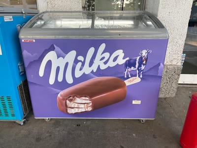 A Milka brand freezer is positioned outdoors on a paved surface. The freezer is purple and features a graphic of an ice cream bar and a cow, with the brand name "Milka" prominently displayed in white lettering. The freezer has a glass lid, and a second blue freezer is partially visible to the left. A red cylindrical object is on the right side of the frame. The background consists of a textured building facade and a reflection of trees and sky on the freezer's glass lid. The scene appears to be in a commercial or retail area in Gorni Dabnik, Bulgaria.