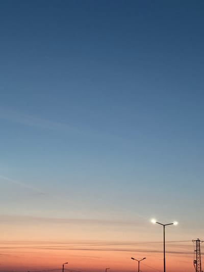 The image displays an outdoor scene at either dawn or dusk in Plodovitovo, Bulgaria. The sky transitions vertically from a deep blue at the top to a lighter blue, then to a pale orange, and finally a vibrant orange-pink at the horizon. Faint linear contrails are discernible in the upper left quadrant. Silhouetted against the lower sky are several utility structures. A prominent street light, positioned in the lower right quadrant, consists of a dark pole supporting two upward-angled arms, each terminating in a brightly illuminated, circular light source. To its left, a smaller, unlit streetlight silhouette is visible. Multiple thin, dark horizontal lines, indicative of power or communication cables, stretch across the mid-ground. On the far right edge of the frame, a section of a dark, metallic lattice power pylon or communication tower is partially visible, extending upwards from the horizon line. No individuals or active subjects are present within the frame.