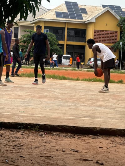 In Amansee, Nigeria, on an outdoor basketball court with a worn, reddish-brown surface, five young men are engaged in a game. One man in a white tank top and black shorts dribbles an orange basketball. Another man in a dark blue t-shirt and black shorts stands nearby, looking towards the basketball player. A third man in a purple and yellow basketball jersey is partially visible on the left. Two other men, one in a black shirt and the other in a blue shirt, stand further back. In the background, a two-story yellow building with solar panels on its roof is visible. Several parked vehicles, including a white van, are also present. The environment appears to be an urban or semi-urban setting.