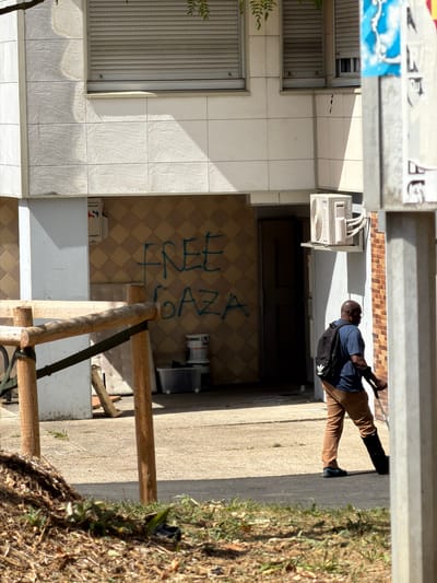 This media file captures an outdoor scene in Évry-Courcouronnes, France. A Black man is walking away from the camera, his back to the viewer. He is wearing a blue t-shirt, brown pants, and a black backpack. He appears to be walking along a concrete path next to a grassy area with dry leaves.
The setting appears to be the exterior of a building with tiled walls and an air conditioning unit visible on the wall. There is graffiti spray-painted in blue on the tiled wall, which appears to say "FREE CIAZ" or similar. Wooden beams and scaffolding are also present in the foreground.
The lighting suggests it is daytime, with shadows cast on the ground, indicating a sunny day. The overall mood is neutral, focusing on the architectural elements and the presence of an individual in an urban or suburban environment. The image's orientation is landscape, but the content is presented rotated 90 degrees clockwise.