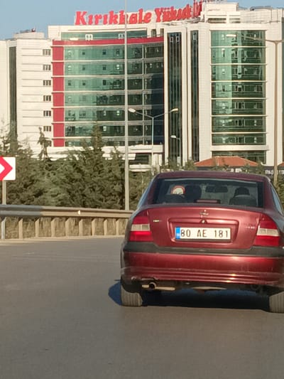 A red sedan with license plate "80 AE 181" is driving on a highway in front of a modern building. The building has a white facade with red accents and green-tinted windows. The text "KIRIKKALE YÜKSEK" is visible in red letters at the top of the building. There are trees and a guardrail along the side of the highway. A directional sign with a white arrow on a red background is visible to the left of the car. The image appears to be taken during the daytime, possibly in the afternoon, given the shadows. The sky is clear and blue. The overall scene depicts a typical urban or suburban highway environment in Turkey.
