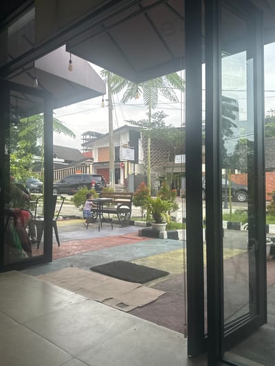 This image captures a view looking out from inside a building, likely a cafe or restaurant, onto a street scene in Sukabumi, Indonesia, on an overcast day. Large black-framed glass doors or windows dominate the foreground, with reflections of the interior, including a ceiling structure, hanging light bulbs, and possibly a person.
Outside, an inviting outdoor seating area is visible with dark metal tables and chairs. One person is seated at a table on the right side of the frame. Potted plants adorn this area, which features a distinctive multi-colored paved ground in shades of blue, yellow, purple, and gray. A black doormat and some lighter-colored floor tiles or mats are also on the ground.
Beyond the seating area, a street is visible with a parked black car. Across the street, several buildings, typical of an urban or semi-urban setting, line the road. One building has a white sign with dark, unreadable text. Lush tropical foliage, including large fern-like plants and other trees, is scattered throughout the scene, contributing to the warm climate aesthetic. The sky is uniformly gray and overcast.