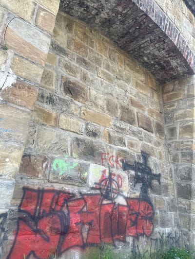 The image depicts a section of a stone archway, likely part of a bridge or viaduct, with visible graffiti on its weathered stone surface. The stonework is rough-hewn and varies in color from light beige to darker brown and grey, indicating age and exposure to the elements. The arch itself is constructed from brickwork, with some damage and debris visible on its underside.

Graffiti is present in several areas. In the lower left quadrant, large red spray paint with black outlines dominates, obscuring much of the stone. Towards the center, red lettering spelling "FCK" is prominent. Above and slightly to the right of the red lettering, a black spray-painted symbol resembling a stylized cross or a winged figure is visible. There are also smaller tags and scribbles in green and blue spray paint scattered across the stone.

The ground beneath the archway is covered in grass and some weeds, with a few small yellow flowers visible on the right side. The lighting suggests it is daytime, with shadows cast by the archway and possibly by an unseen object. The overall impression is one of urban decay and the transient nature of public spaces marked by graffiti.

This scene is likely located in Wuppertal, Germany, specifically on the historic Sonnborner Viadukt, a railway viaduct known for its extensive stonework and frequent graffiti, which is a recurring issue managed by local authorities.