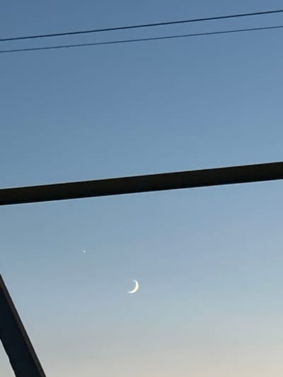 This image depicts a celestial view of the sky over Sibay, Russia. Two prominent celestial bodies are visible: a crescent moon and a bright star or planet. The moon is positioned to the right, showing its characteristic curved shape. To the left of the moon, a smaller, luminous point, likely a planet or star, is visible.

The sky is a gradient of blue, transitioning from a deeper blue at the top to a lighter shade near the horizon. Two dark, thin lines, possibly power lines or cables, stretch horizontally across the upper portion of the frame, suggesting an urban or semi-urban environment. A thick, dark, angled object, possibly part of a structure or scaffolding, occupies the lower left portion of the image, partially obscuring the sky. The overall lighting suggests either dawn or dusk.