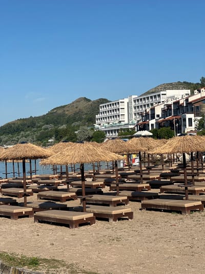 The image depicts a beach in Topola, Bulgaria, featuring rows of empty sun loungers and thatched umbrellas set up on a sandy shore. In the background, a modern, multi-story hotel complex with white facades and balconies overlooks the sea. Lush green hills and trees form a backdrop behind the hotel. The sky is clear and blue, indicating a sunny day. The beach appears to be prepared for visitors, with the loungers neatly arranged and ready for use, though no people are visible in the scene. The overall atmosphere is serene and suggests a tourist-oriented location.