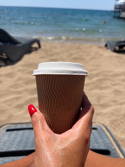 A close-up, side-view shot shows a person's hand holding a brown, corrugated paper coffee cup. The cup has a white rim and lid. The hand has bright red nail polish on one fingernail, visible on the right side of the frame. The background is a sandy beach with the gentle waves of the ocean on the left. The sand shows ripples, suggesting it may have been recently disturbed or wind-blown. In the shallow water, there are some reflections from the sunlight, creating bokeh effects. In the mid-ground, blurred dark shapes of beach chairs are visible. The scene is bright and sunny, implying a daytime setting, likely warm weather suitable for a beach outing. The overall impression is one of relaxation and enjoying a beverage in a coastal environment. The location context provided is Trikomo, Northern Cyprus, which aligns with the beach and sea setting. No visible text is present on the cup or in the scene.