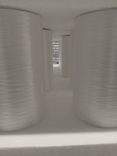 The image shows a perspective view of several large, white, cylindrical spools of material, possibly yarn or fiber, arranged in a row. The spools are tightly wound and have a textured surface. They are positioned within a large, brightly lit, indoor space with a white, textured ceiling and floor. In the background, a section of a brick wall is visible, along with what appears to be a shelf or partition. The overall impression is one of an industrial or storage environment. This image was taken in Deurne, Netherlands, likely within a manufacturing or warehousing facility. There are no people or animals visible in the image.