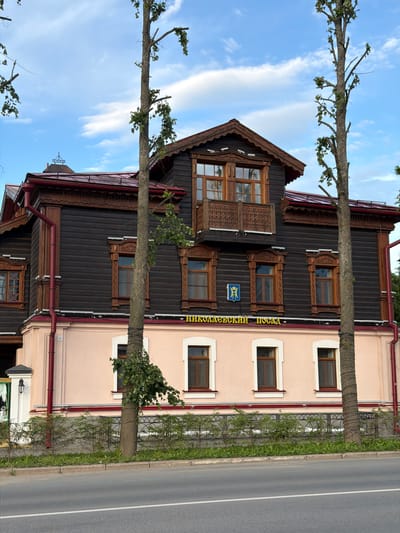 This image shows the Nikolayevsky Posad in Suzdal, Russia, a two-story building with a dark wooden upper floor and a light pink lower floor. The building features intricate wooden carvings around the windows and a balcony with ornate railings. A prominent tree with sparse foliage stands in front of the building, with its trunk wrapped in decorative lights. In the background, a blue sky with scattered clouds is visible. A street runs in front of the building, with a sidewalk and a decorative metal fence separating the property from the road. The scene is captured during daylight, under a slightly overcast sky. There are no people or animals visible in the image.