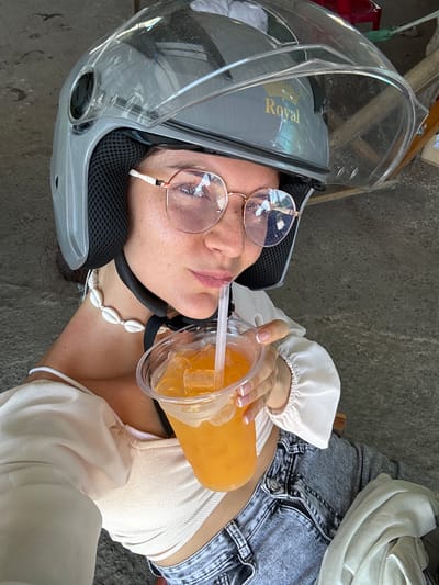 A young woman wearing a grey helmet and glasses is taking a selfie while holding a plastic cup of orange juice with ice and a straw. She is also wearing a white top and denim shorts. The background appears to be an outdoor setting with a concrete ground and some blurred objects in the distance, possibly a street scene in Hồng Sơn, Vietnam.