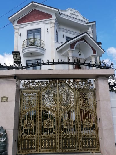 The image shows the ornate, gold-colored double gates of a property, likely a residence, in the city of Lam Vien - Da Lat, Vietnam. The gates are intricately designed with detailed carvings and emblems, featuring what appear to be coats of arms on each panel. The gates are set within a beige stone wall that frames the entrance. Above the gates, a modern two-story house is visible. This house is white with red brick accents on the upper level and a black tiled roof. A balcony with a curved railing extends from the second floor, and a prominent outdoor light fixture is mounted on a pole to the left of the house. The sky above is a clear blue with a few scattered white clouds. The overall impression is one of wealth and elaborate architectural style.