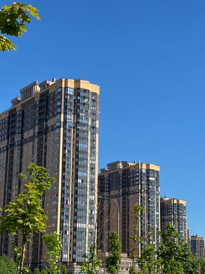 In Pargolovo, Russia, the image captures a clear, bright, blue sky above several modern, tall residential buildings. The buildings are characterized by their beige and brown facades, with numerous windows and balconies. Lush green trees and foliage are in the foreground and middle ground, partially obscuring the lower sections of the buildings. The architecture suggests a contemporary urban or suburban setting. No people are visible in the scene.