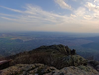 The image depicts a high-altitude view overlooking a city, likely Ichera, Bulgaria, given the prompt. The foreground is dominated by rocky terrain and sparse vegetation, including leafless shrubs. The city stretches across the midground, with discernible urban structures and a somewhat hazy atmosphere. In the distance, the sky is blue with scattered clouds. No people or animals are directly visible. An unidentified weathered, possibly metallic structure is partially visible at the lower left edge. The overall setting suggests a mountain peak vantage point.