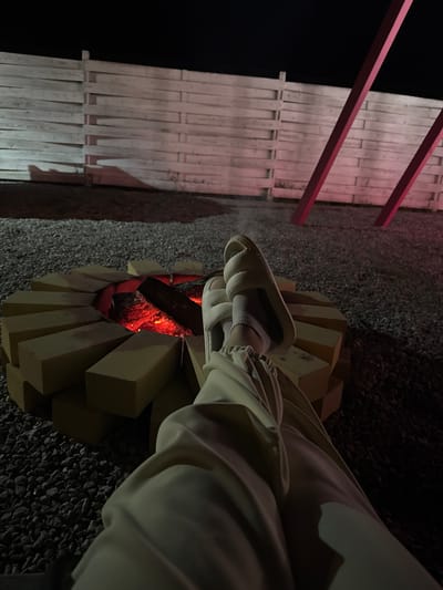 The image shows a person sitting by a fire pit. The fire pit is constructed of light-colored rectangular blocks arranged in a circle. There is a fire burning inside, with visible flames and burning wood. The person is wearing light-colored pants and sandals with socks. The background shows a wooden fence-like structure with red supports, and a dark gravel surface. The location is likely outdoors at night. The city of Pad', Russia is not discernible from the image.