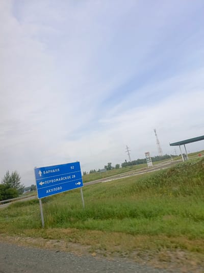 A blue road sign with white lettering is visible in the image. The sign indicates directions to "Barnaul" (92 km), "Pervomayskoye" (28 km) to the left, and "Akulovo" (1 km) to the right. The sign is supported by two metal poles. The background shows a cloudy sky, a gas station with a price board, cell towers, power lines, and a grassy field with scattered trees and wildflowers. The ground in the foreground is a gravel road. The scene appears to be a roadside location in Vidonovo, Russia.