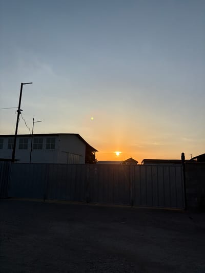 A vertical, low-angle, full shot captures a sunset scene behind a metal fence and industrial buildings in Dolni Dabnik, Bulgaria. The sky transitions from a pale blue at the top to a warm orange glow near the horizon. The sun, a bright orb, is partially visible, casting light rays and creating a lens flare effect on the upper right side of the image. A dark, silhouette of a building with multiple windows is on the left, with a utility pole and wires extending upwards. A tall, gray corrugated metal fence runs horizontally across the foreground, obscuring the lower parts of the buildings and the ground. The ground in front of the fence appears to be gravel or dirt. Several other building structures, also in silhouette, are visible beyond the fence.