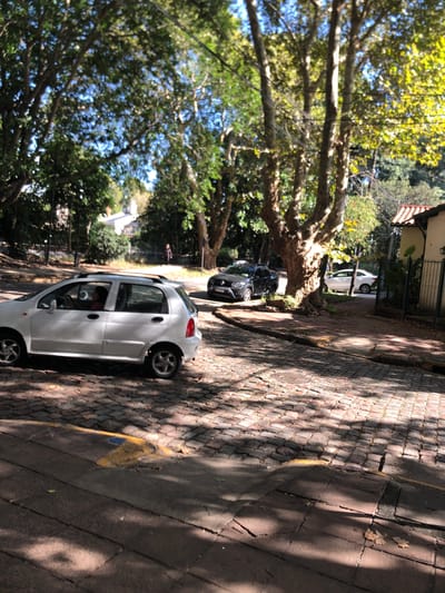 The image shows a street scene in Martínez, Argentina. A white car is parked on the right side of the cobblestone street, with its rear facing the camera. The street is lined with trees providing shade and creating shadows on the pavement. Further down the street, at least one other car is parked. There is a curb separating the street from the sidewalk. The presence of trees suggests a residential area.
