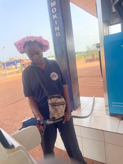 A person with a pink head covering is holding a blue fuel pump nozzle at a gas station. They are wearing a dark blue shirt with a circular logo that includes text "WEST SIDE PETROCHEMICALS LTD" and "ENERGY & ENGINEERING." The person has a purse with a patterned design slung over their shoulder. The gas station has a blue column with the word "SMOKING" printed vertically in reverse. The background shows a typical Oturkpo, Nigeria environment.
