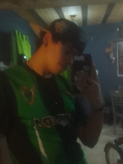 A person takes a selfie indoors, wearing a green and black shirt with a shield-shaped logo and a baseball cap with a design on it. They hold a phone with a decorated case in their hand. The background is a room with blue walls, a green curtain, and a ceiling with exposed beams. A chair and a picture frame are visible in the background. The lighting is somewhat dim.