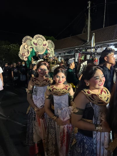 The image captures a nighttime cultural procession on a street in Munggu, Indonesia. In the foreground, three Balinese women are presented in elaborate traditional attire and theatrical makeup. Their faces exhibit light foundation, flushed cheeks, and defined eyebrows. The two women on the left directly face the viewer with slight smiles, while the woman on the right smiles and looks to her right. Each woman wears a golden, ornate headdress, a large golden neck collar with red and black accents, and golden armbands and bracelets. Their lower garments are patterned sarongs; the two central women have white fabric draped over their chests, with visible red staining on the lower portion of their sarongs. The second woman from the left holds a rectangular object, possibly a book or tablet, displaying a drawing of a hand.
Behind these women, a crowd of individuals, primarily men in traditional Balinese black shirts, udeng headwear, and sarongs, extends down the street. In the midground, a large, ornate ceremonial float or structure, predominantly white and gold with green drapery, is visible, carrying several participants. The background consists of a dark night sky, a building with a brown tiled roof, utility poles, and overhead power lines. A partial sign displaying "APDAH QC" is visible on a building facade. Artificial lighting illuminates the scene, indicating an evening event.